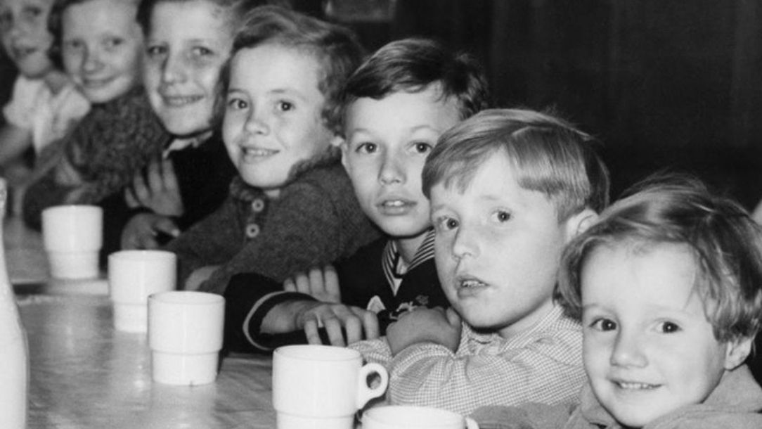 A black and white photograph showing small children sitting down at a table waiting for jelly and cake.