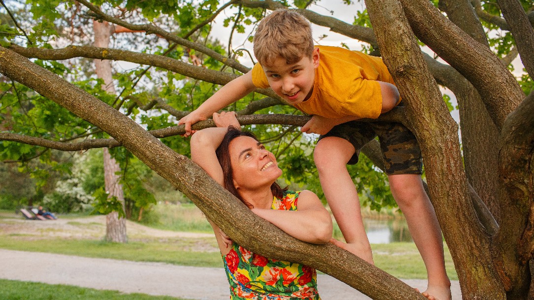 A boy wearing a yellow t shirt and brown shorts faces the camera while climbing a tree. His mother, stood on the ground and wearing a floral t shirt looking up at him smiling