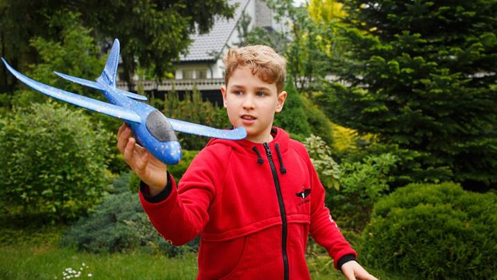 A boy wearing a red zip-up fleece stands in a garden holding a toy plane up in the air