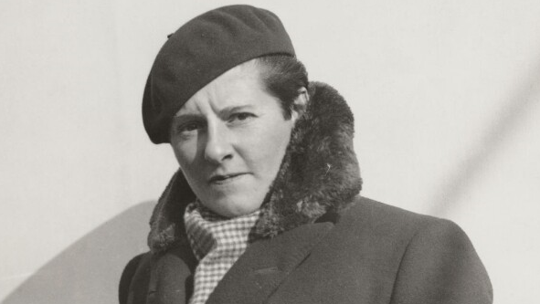 A black and white image of Joe Carstairs, who is wearing a dark beret, checked scarf and dark coat with a fluffy trim.