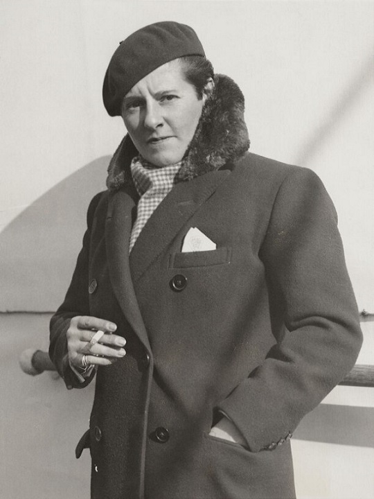 Joe Carstairs A black and white photo of Joe, who has short hair and is holding a cigarette while looking at the camera. She's wearing a dark coat with a fluffy trim, dark beret and checked scarf.