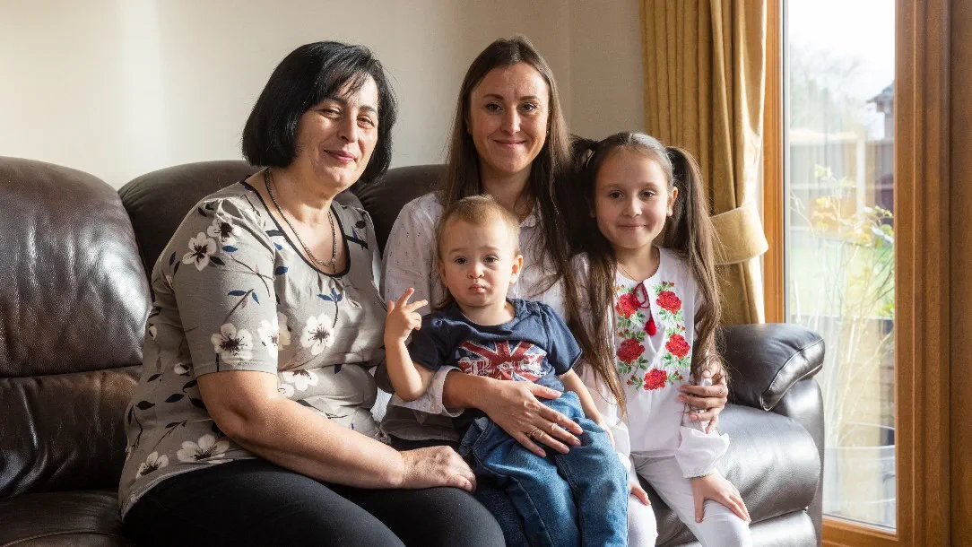 Two women and two children sit on a sofa smiling at the camera. They have been living in the UK since arriving from Ukraine in September 2022
