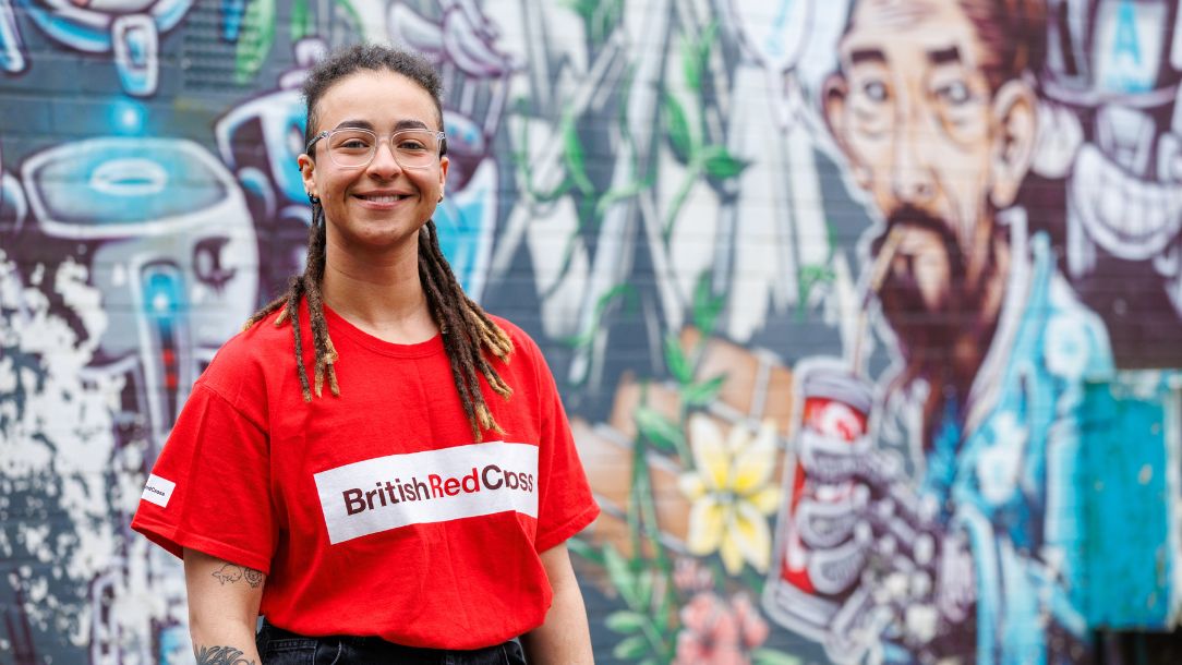 Kat, a British Red Cross volunteer, wears a British Red Cross t-shirt and stands against a colourful wall of graffiti.