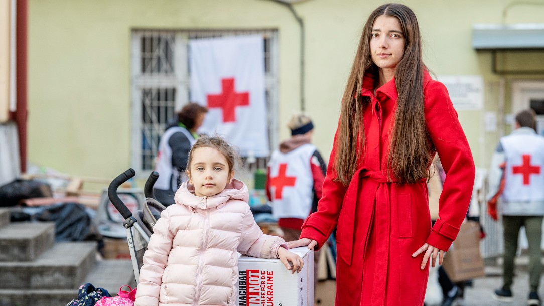 A woman and child look at the camera standing next to the hygiene kit they have just received. Two volunteers wearing Red Cross uniforms are seen in the background
