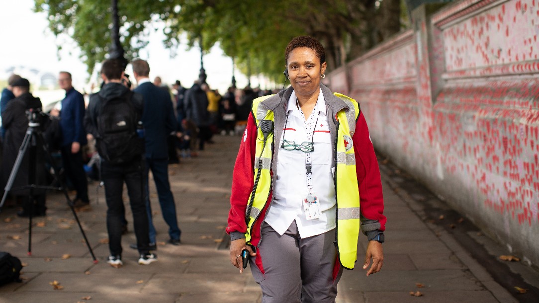 British Red Cross volunteer Kofo walks toward the camera in her Red Cross uniform, in front of a background of people queuing along the River Thames to see Her Majesty the Queen