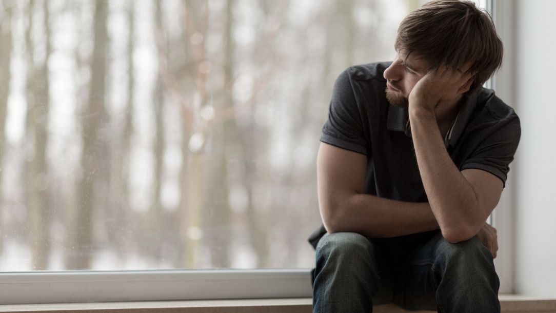 Lonely young man gazes out of a window with his head in his hands.