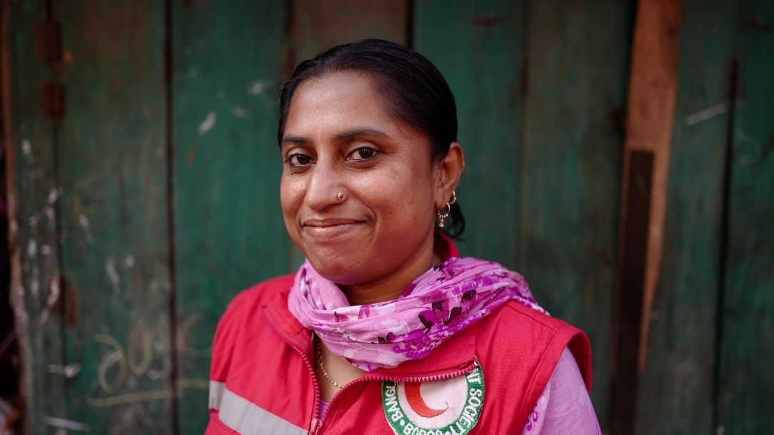 A photograph of a woman in Bangladesh wearing a Red Crescent vest and pink scarf, smiling at the camera.