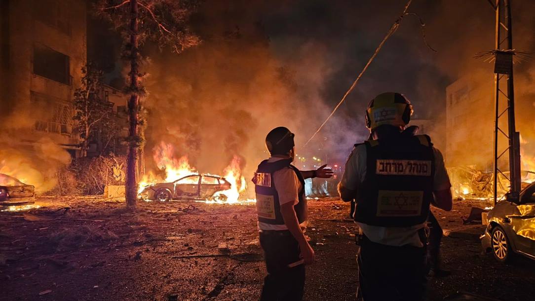 A Magen David Adom rescue worker with a burning car in the background.