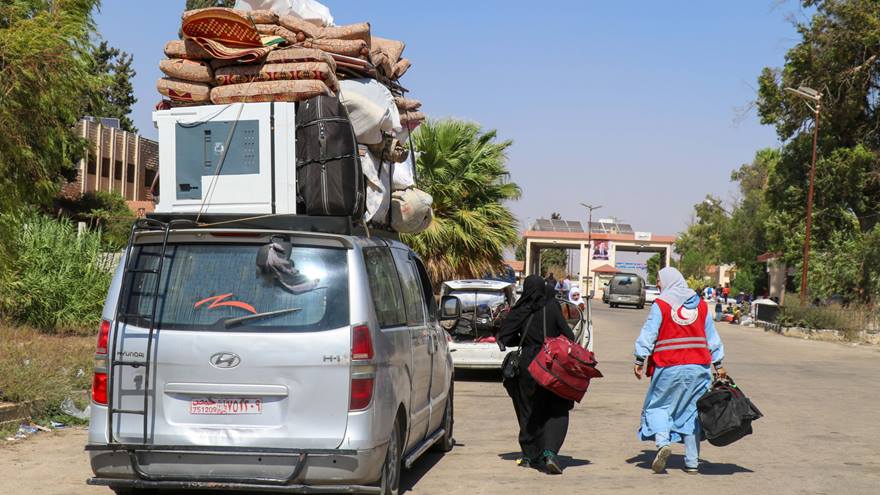 A Red Crescent worker walks with a woman, walking next to a car with belongings on the roof.