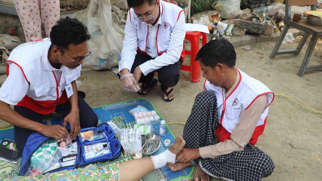 A member of the Myanmar Red Cross Society wearing a vest bearing the red cross emblem applies a bandage to the foot of an injured person, who is lying on the floor