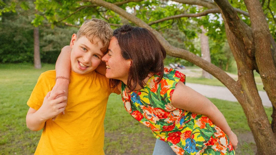A mother and son pose for the camera in the park with their arm around each other. The son is wearing a yellow t-shirt, while his mother is wearing a floral top. 