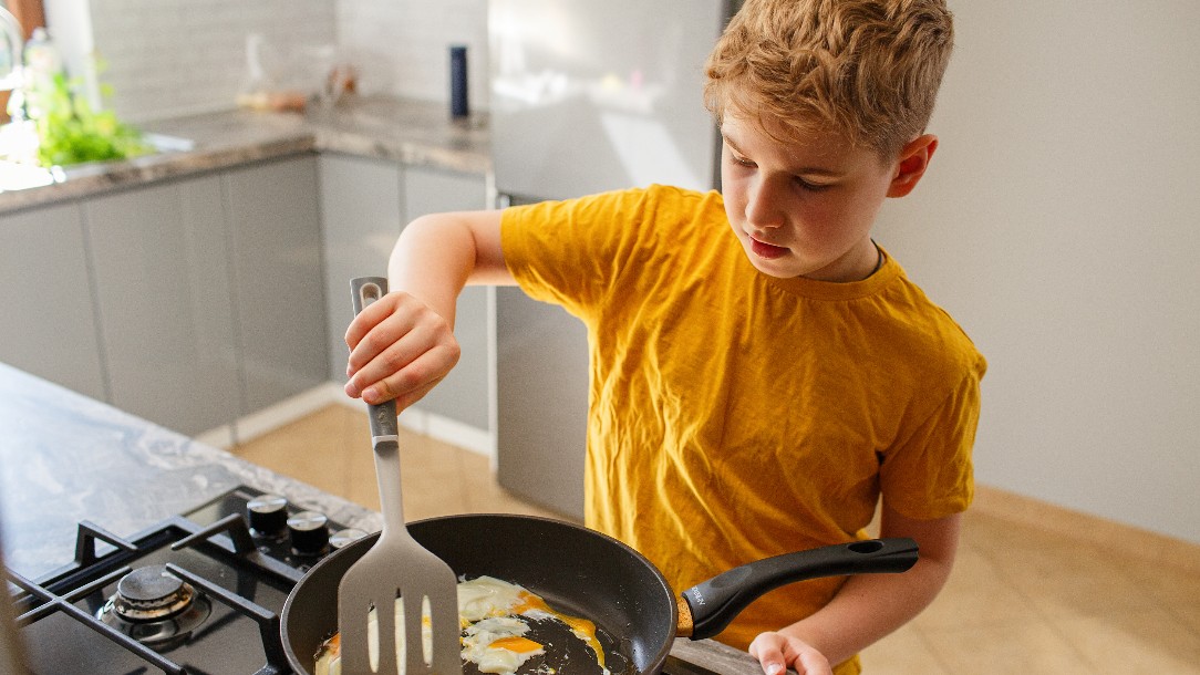 A boy wearing a yellow t-shirt fries an egg in a frying pan