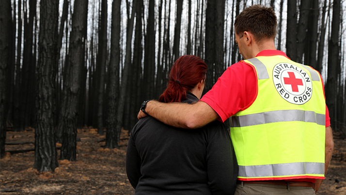 Standing in front of woods blackened by fire with their backs to the camera, an Australian Red Cross volunteer puts his arm around a woman as she lowers her head.