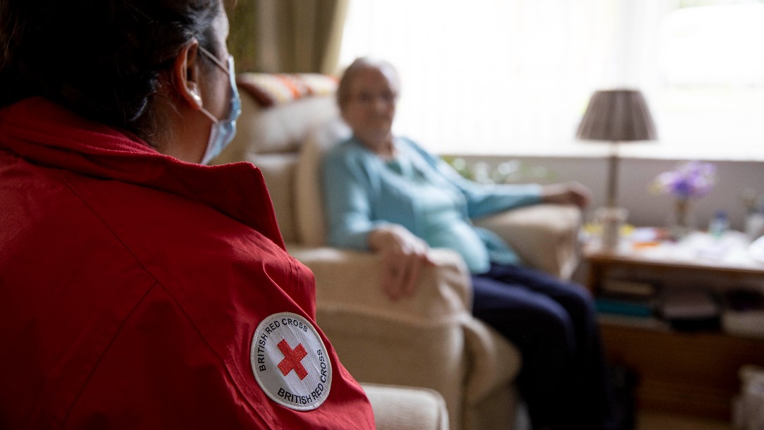 Natalie sits, wearing her British Red Cross uniform and a face mask, opposite Eunice during a visit to her house