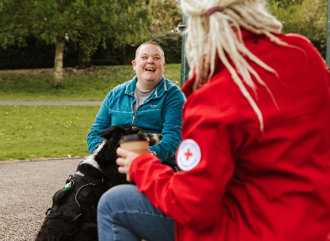 Red Cross service user Lisa laughs with community connector Tracey on a park bench. Lisa's two dogs sit at their feet.