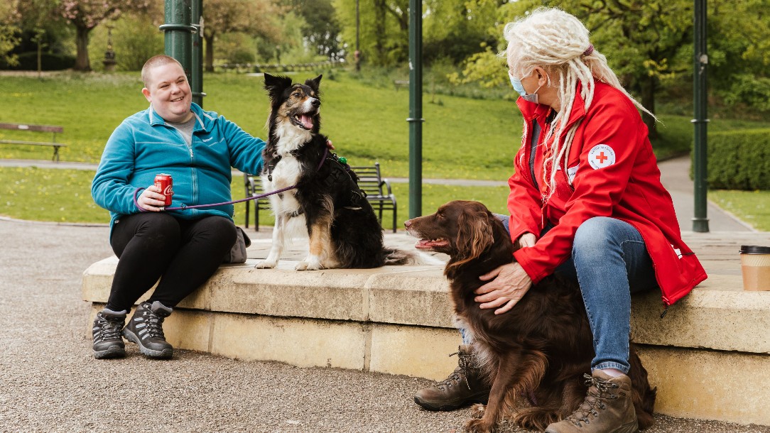 Lisa and Tracey sit on a wall in the park, with Tracey's dogs in between them. Tracey is wearing her Red Cross uniform and a face mask and Lisa is smiling