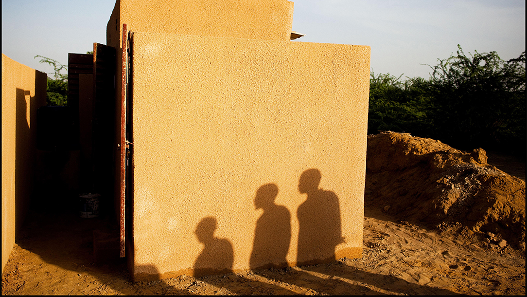 In Niger, shadows of three people show against the brown mud wall of a house. 