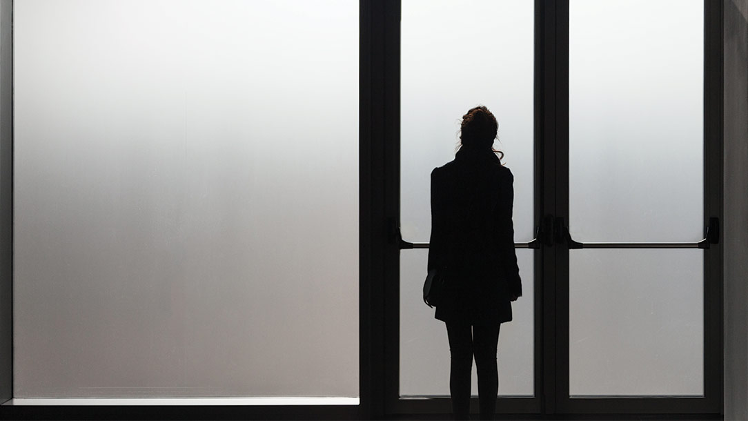 A young woman stands in shadow at a glass wall in a public building. 