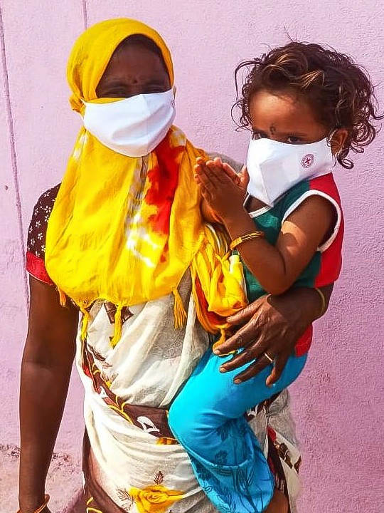 A woman in Telangana, India, holds her child on her hip. They are both wearing face masks. 