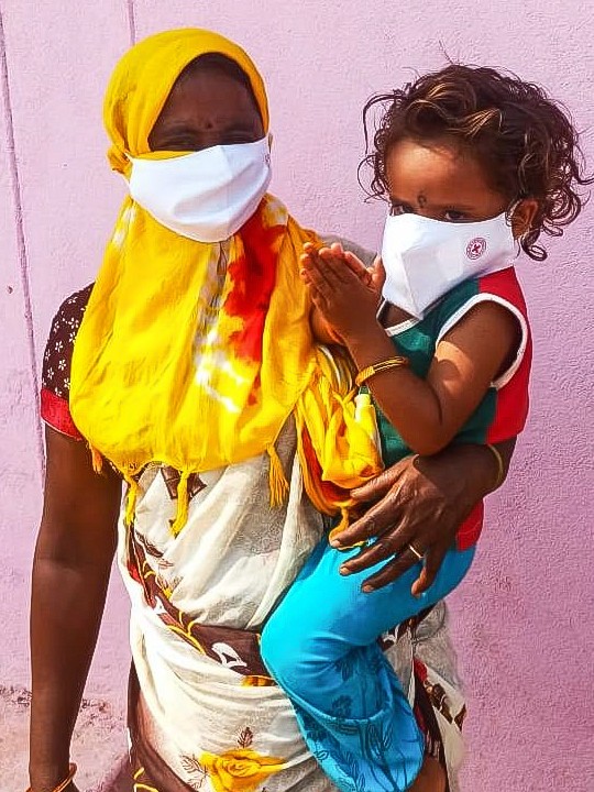 A woman in Telangana, India, holds her child on her hip. They are both wearing face masks.