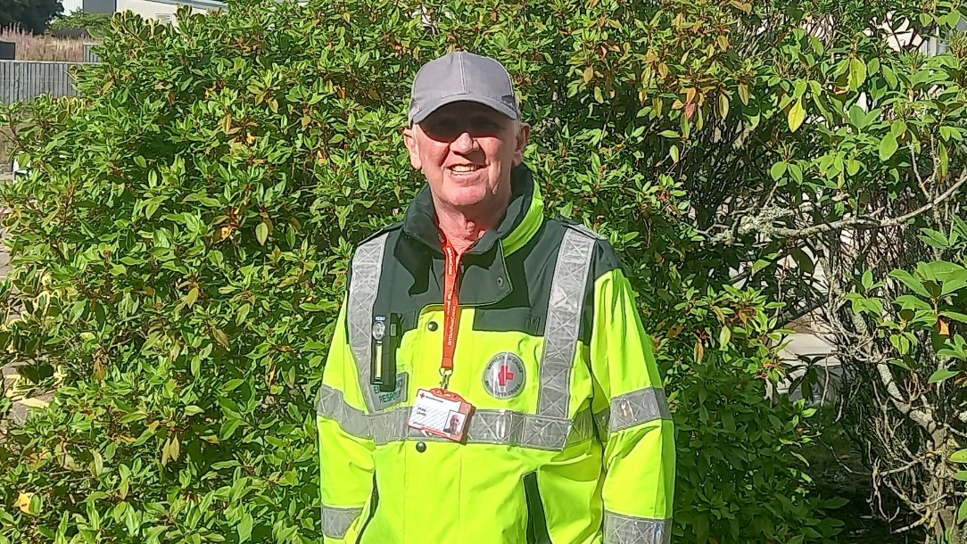 A British Red Cross volunteer wearing a high vis jacket smiles at the camera, stood in front of a bush