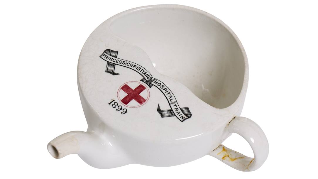 White ceramic cup with teapot-like funnel, small handle and half covered in ceramic. On the ceramic covering the cup, there's a banner printed that says Princess Christians Hospital Train, a red cross and the numbers 1899