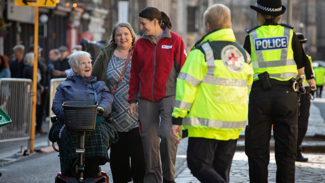Two women, one in a British Red Cross uniform, walk along with a woman who is on a mobility scooter. A police officer and a fellow Red Cross volunteer are seen in the foreground