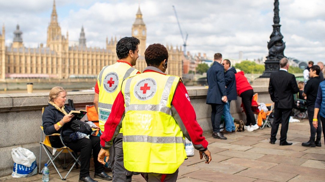 Two British Red Cross volunteers in uniform walk away from the camera, in front of a queue of people waiting to see the Queen. The Palace of Westminster is in the background.