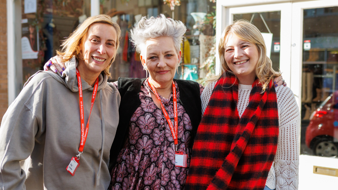 Three volunteers wearing Red Cross lanyards smile in front of the Penarth Red Cross charity shop 