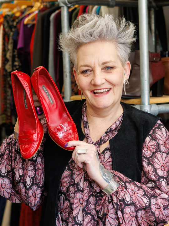 Charity shop volunteer Rachel smiles at the camera holding up a pair of red shoes