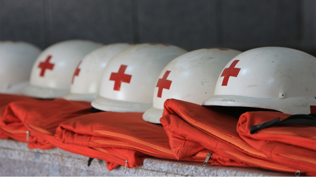 A row of Red Cross helmets and folded emergency response uniforms sit on a shelf.