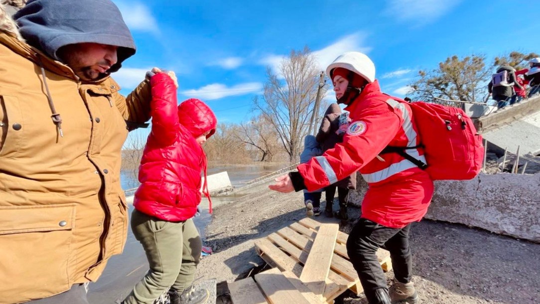 Red Cross volunteer supports man and young child at river crossing in Demydiv