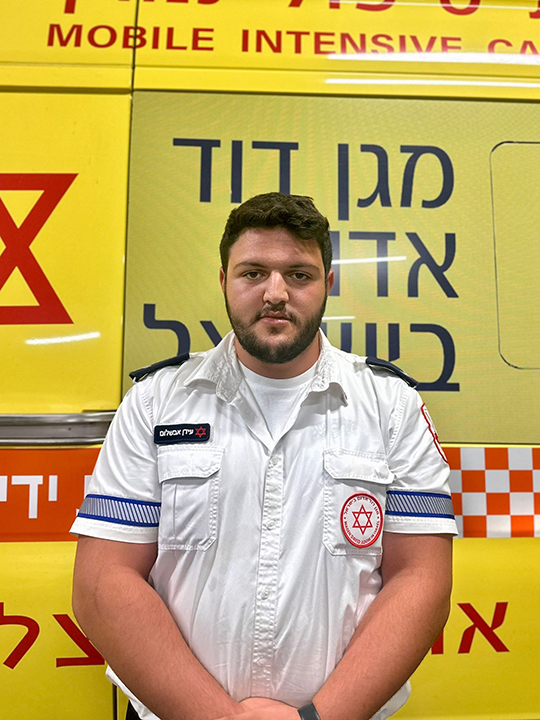 A man wearing a Magen David Adom uniform stands looking to camera, with an ambulance parked behind him