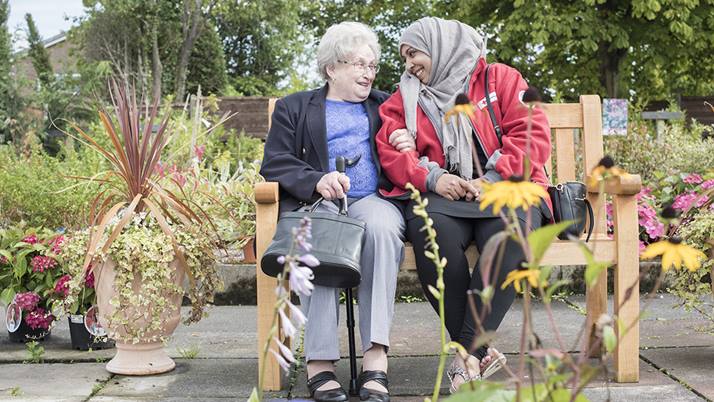 A British Red Cross volunteer sits arm-in-arm on a bench in a garden with an older woman who was feeling lonely as the two smile at each other.