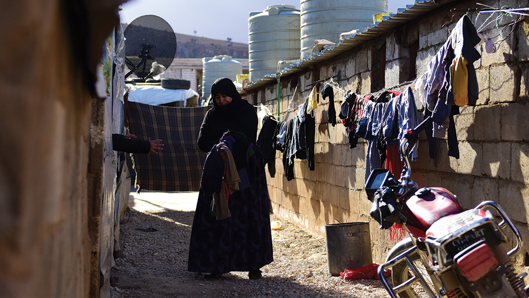 In Syria, a woman walks in a alley between houses collecting laundry that is hanging along the wall of one house.