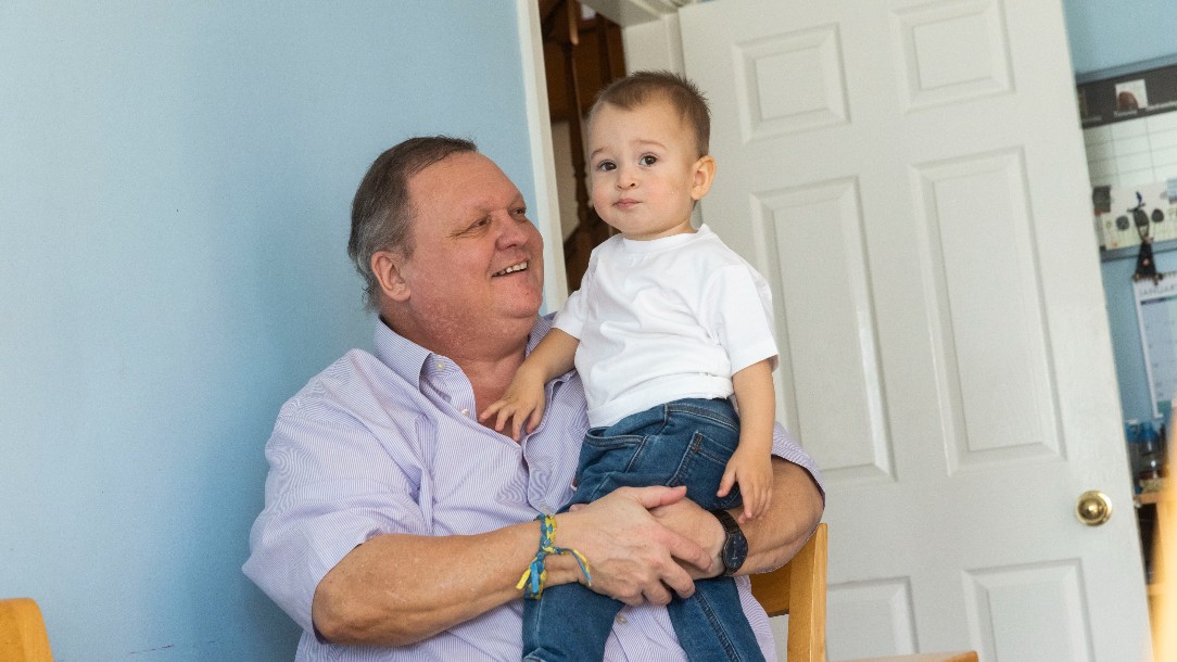 A man, Tony, sits on a chair in his kitchen, smiling at Makar, an 18 month old boy who moved to the UK from Ukraine with his family in 2022