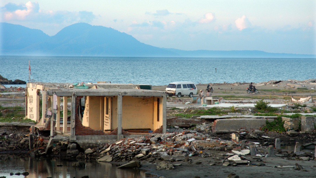 Image of a badly damaged building surrounded by debris in the aftermath of the Indian Ocean Disaster. Open water is seen in the backgrounds, with mountains in the horizon