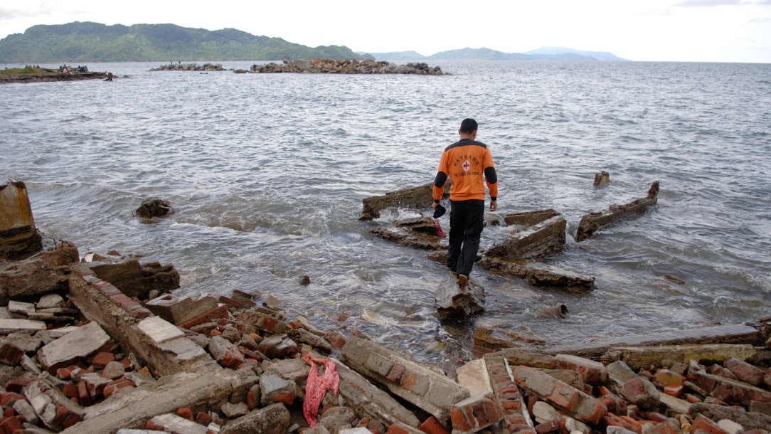 A man looks out to sea in the aftermath of the 2004 Indian Ocean tsunami. In the foreground there is the rubble of buildings damaged by the disaster