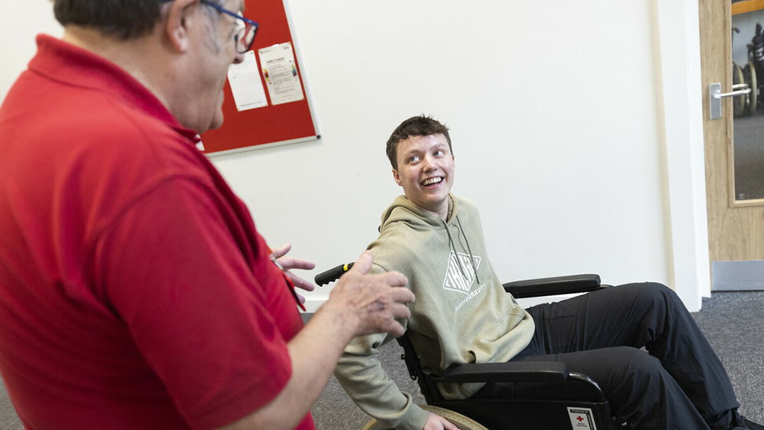 A British Red Cross volunteer and a man in a wheelchair laugh together in the countryside.