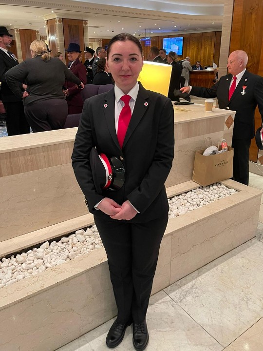 In this full-length photo, volunteer Irina faces the camera wearing her British Red Cross ceremonial uniform, which consists of a grey suit and a red tie