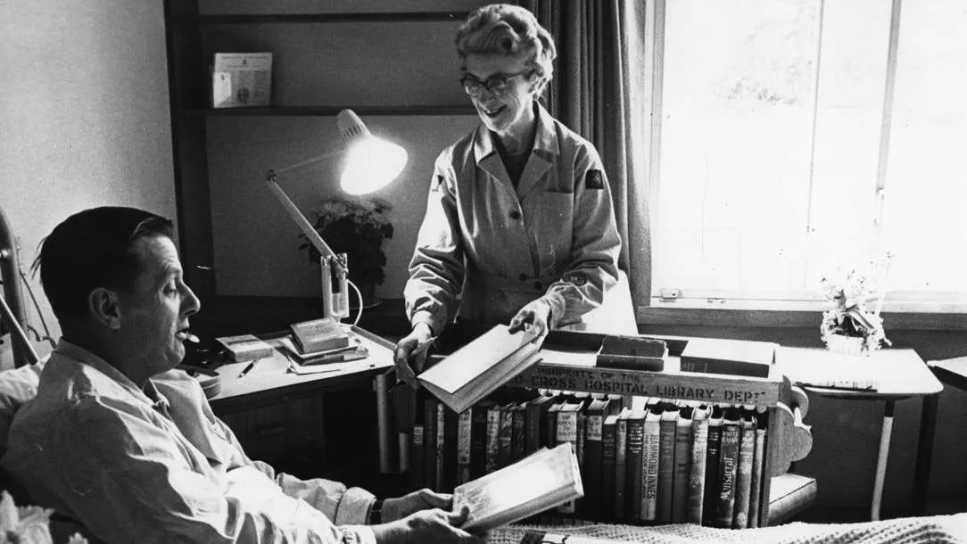 Black and white photo of a British Red Cross nurse handing a man a book.