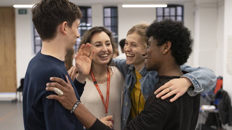 Four young people, two boys and two girls, hugging and laughing in a large theatre rehearsal room