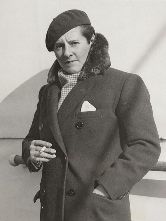 A black and white photo of Joe, who has short hair and is holding a cigarette while looking at the camera. She's wearing a dark coat with a fluffy trim, dark beret and checked scarf.