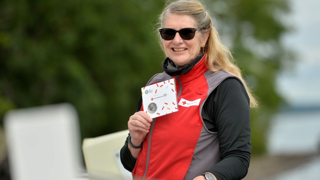 Scottish volunteer, Linda, is dressed in British Red Cross uniform and holds up a British Red Cross 150th anniversary commemorative coin for her work on the support line during the pandemic.