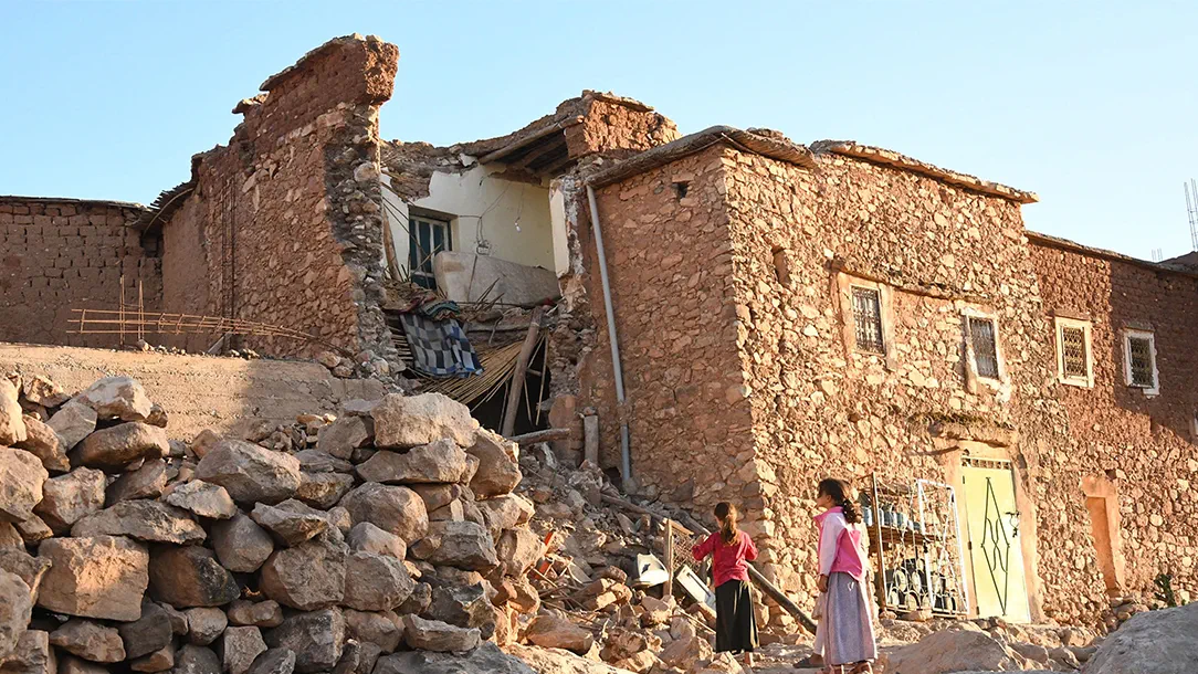 Picture of a building damaged by the earthquake in Morocco, originally made of stones, with two girls standing in front of a pile of rubble.