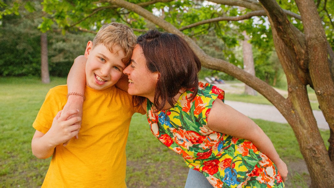 A mother and son pose for the camera in the park with their arm around each other. The son is wearing a yellow t-shirt, while his mother is wearing a floral top.