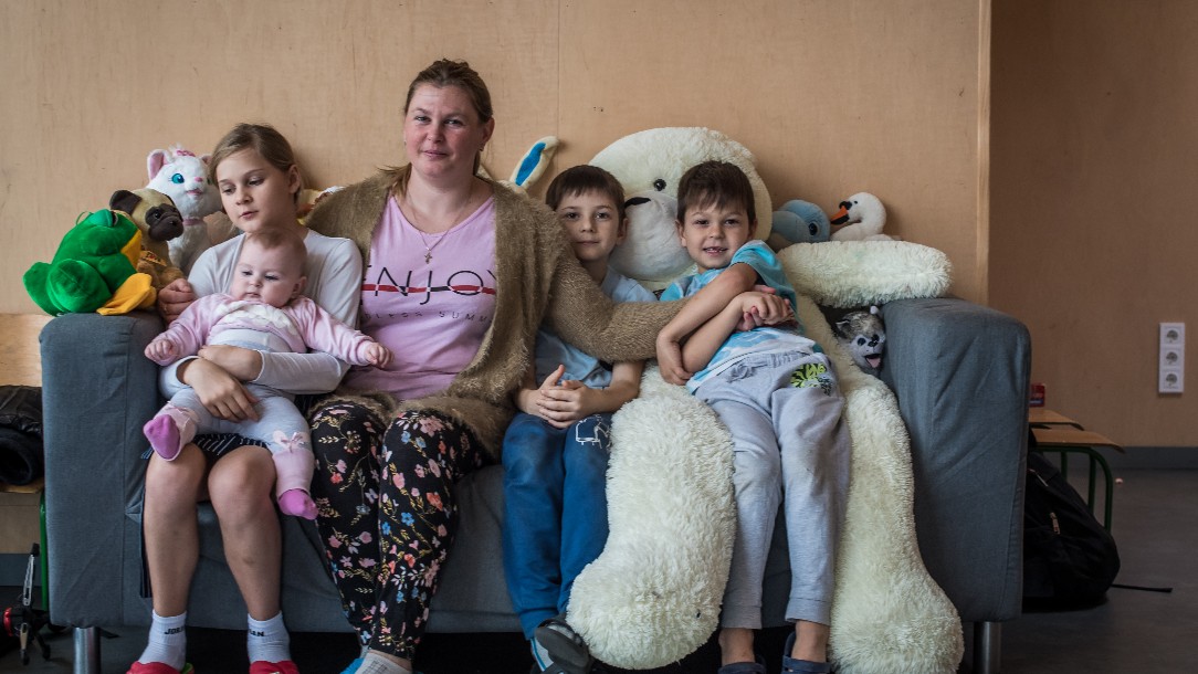 Olha and and her four children sit on a sofa smiling at the camera. One of the children is sitting on a big teddy bear
