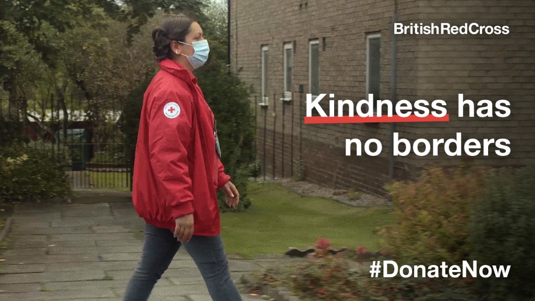 A still from a British Red Cross video shows a British Red Cross volunteer walking along a road, wearing her uniform and a face mask