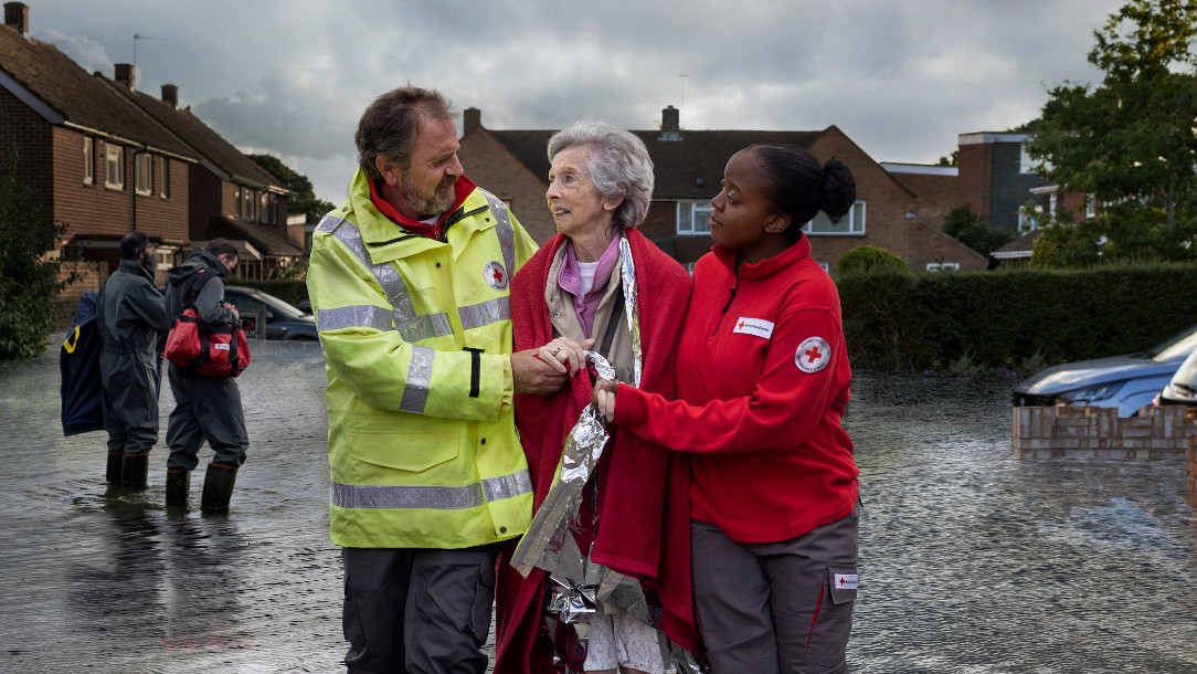 Two British Red Cross volunteers help a woman whose home has been flooded. She is wrapped in a blanket as they wade through flood water together.